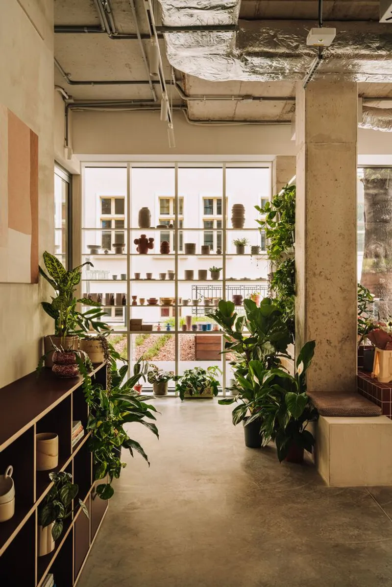 View through planted interior toward white metal display shelving framed by concrete columns and exposed ceiling services