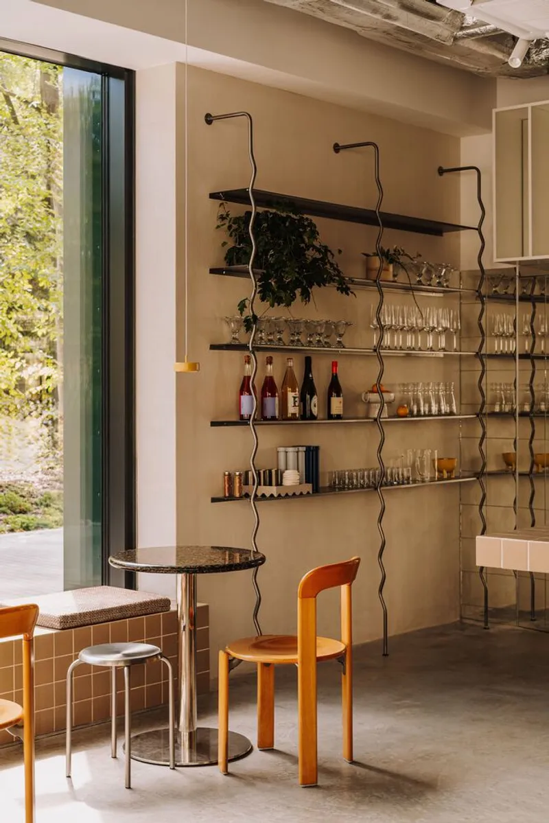 Wavy metal shelving with bottles and glassware beside a small round table and orange chair