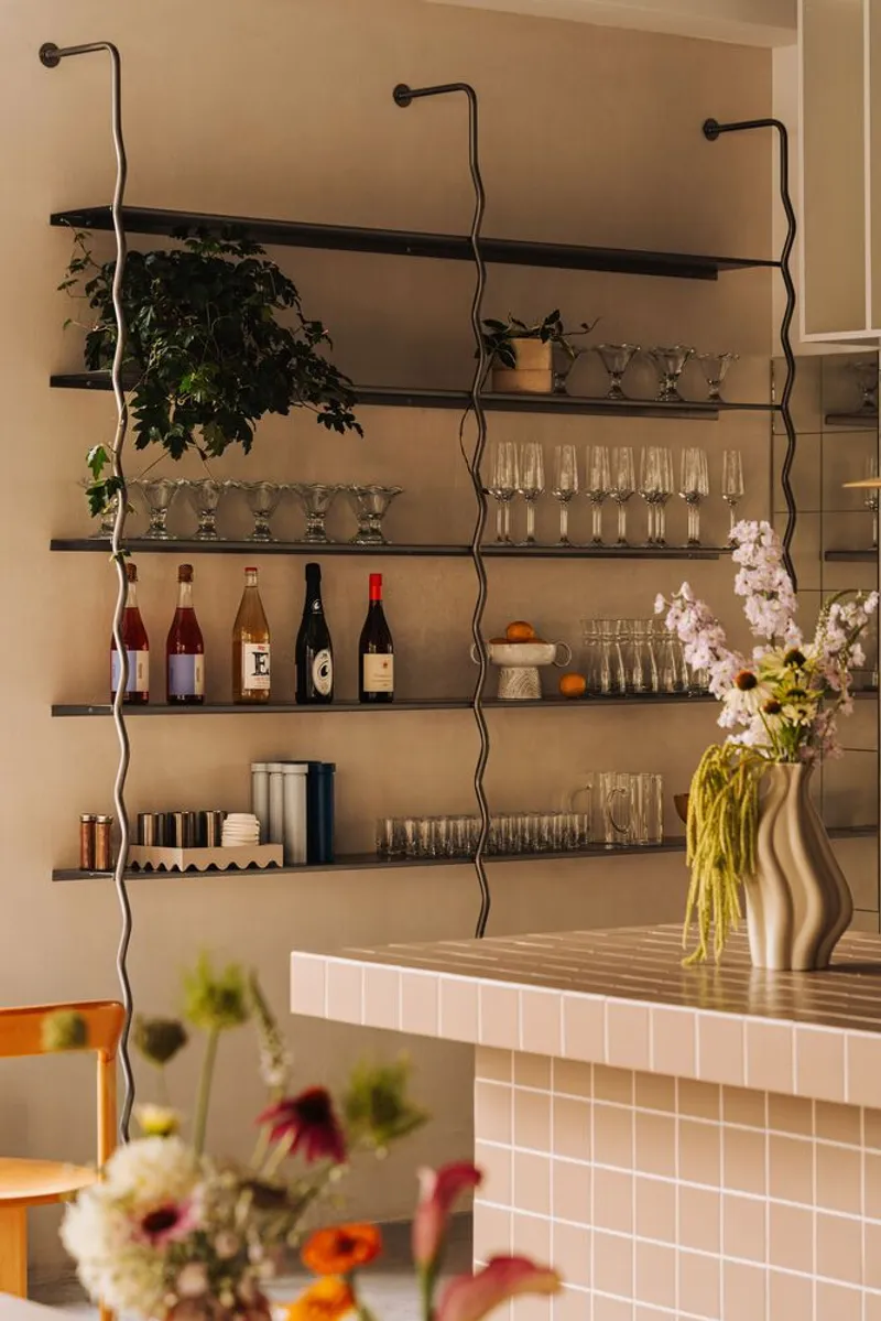 Wall-mounted shelving with wavy metal supports displaying wine bottles, glassware and potted plants