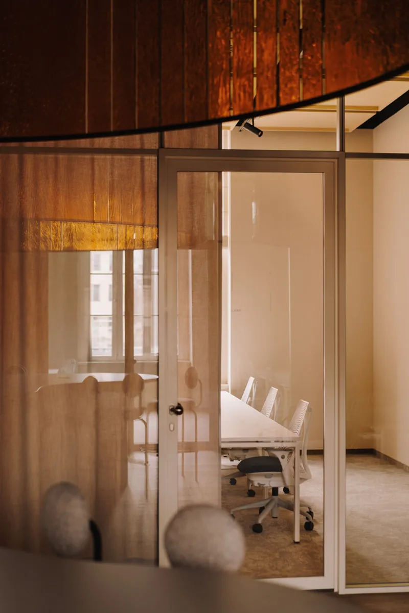 View through glass partition to meeting room with chairs beneath sheer curtain and pendant