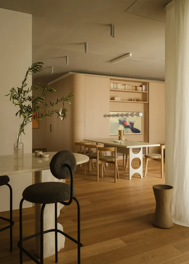 Dining area with oak table and chairs beside a potted tree and sheer curtain