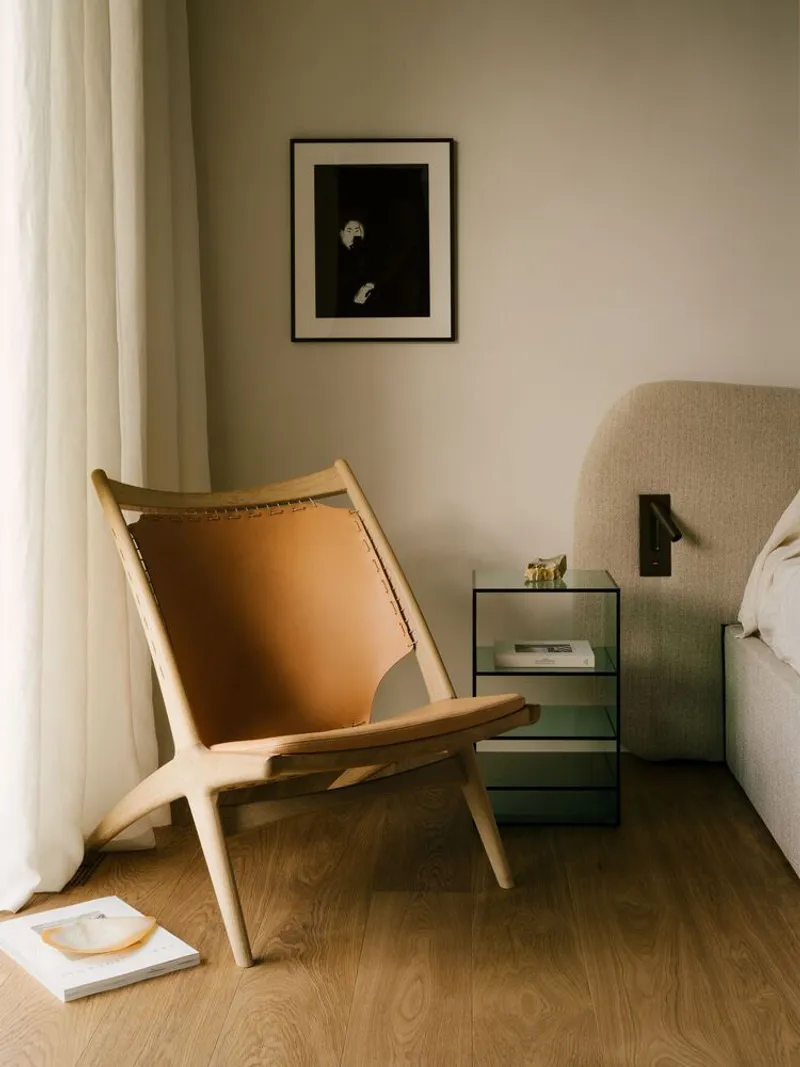 Timber lounge chair with leather sling seat beside a glass bedside table and draped curtain