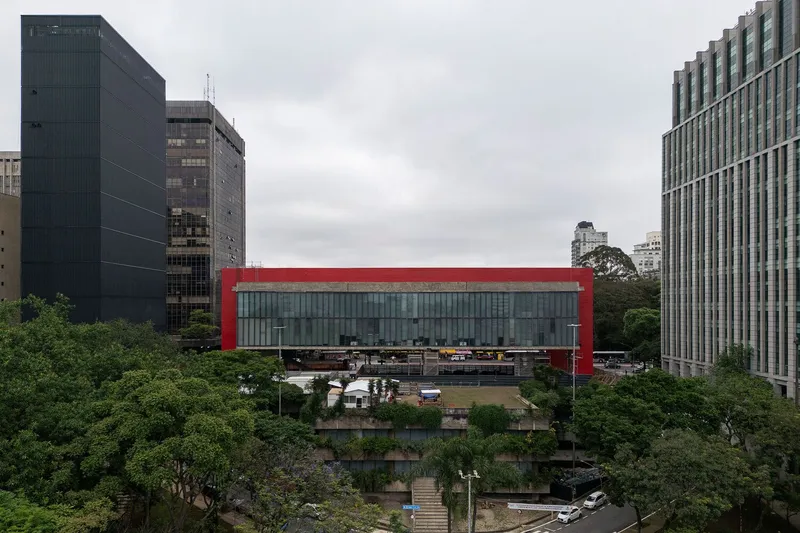 Red-framed glass bridge structure spanning between buildings above a planted sunken plaza on an overcast day