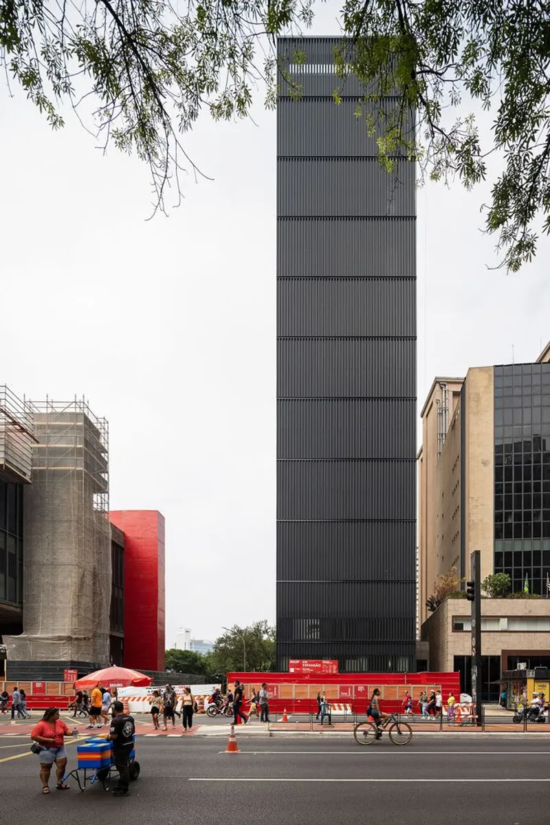 Tall vertical tower with ribbed metal cladding rising above pedestrians and red temporary barriers along the street