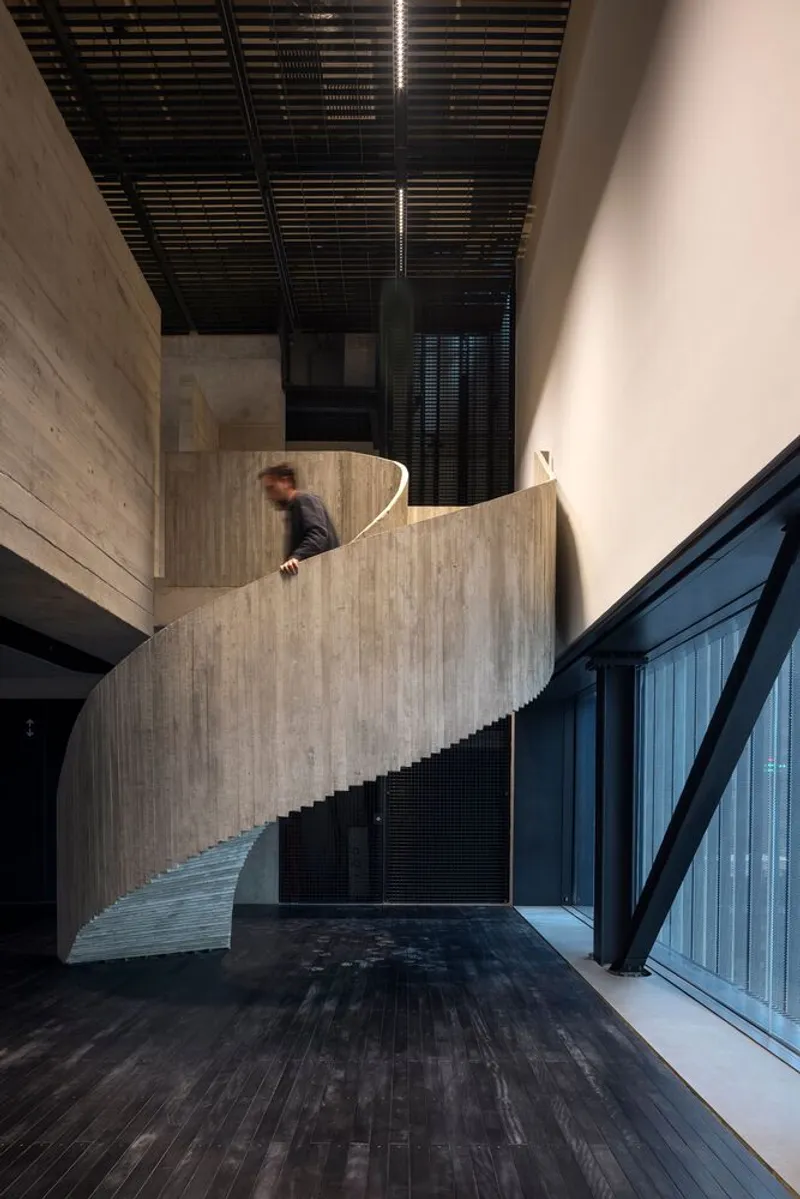 Curving board-formed concrete staircase rising through a double-height lobby with black slatted ceiling and a figure ascending