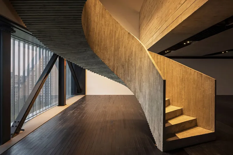 Curved timber staircase wrapping around a board-formed concrete wall under a black slatted ceiling