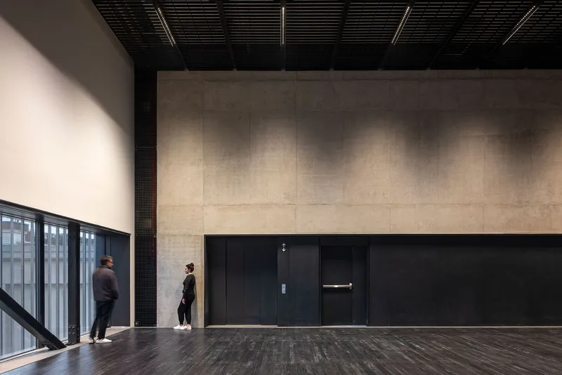Interior lobby with dark timber floor, black tiled wall, and two figures near the elevator