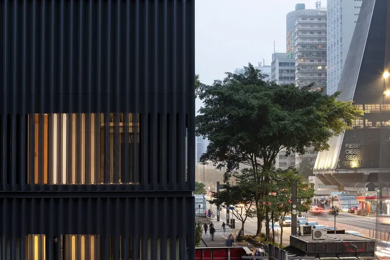 Black corrugated facade with vertical window openings illuminated at dusk beside street trees and traffic