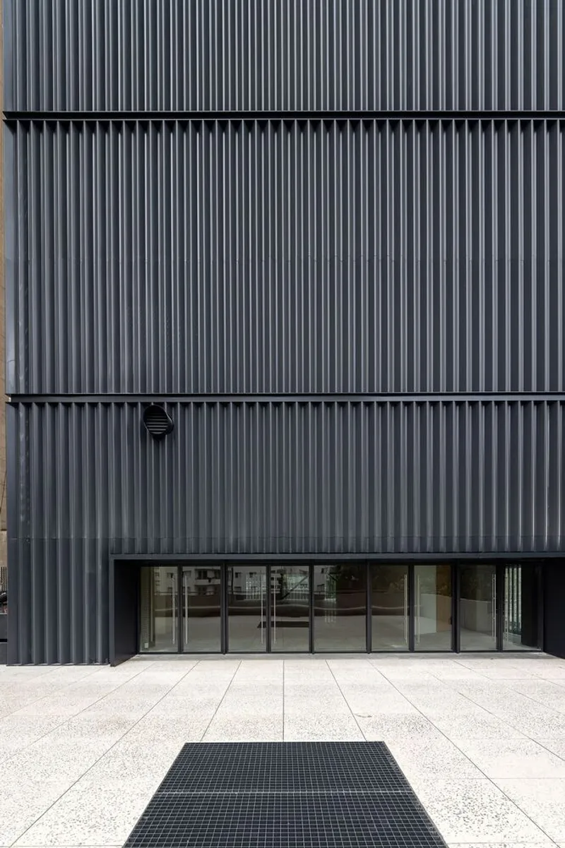 Street-level entrance with glass doors set into a black corrugated metal facade and concrete pavement