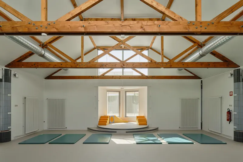Multipurpose hall with exposed timber trusses, exercise mats on the floor and a raised platform below symmetrical windows