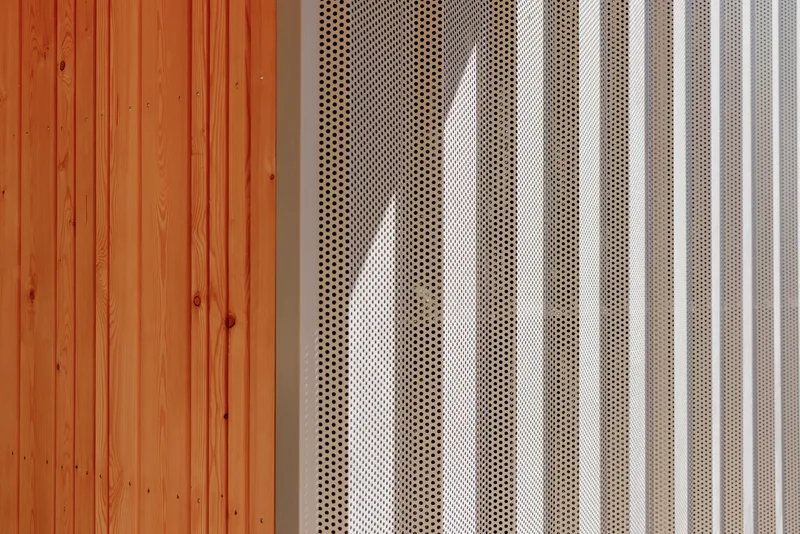 Detail of vertical timber cladding meeting perforated metal screening with sunlight filtering through the pattern