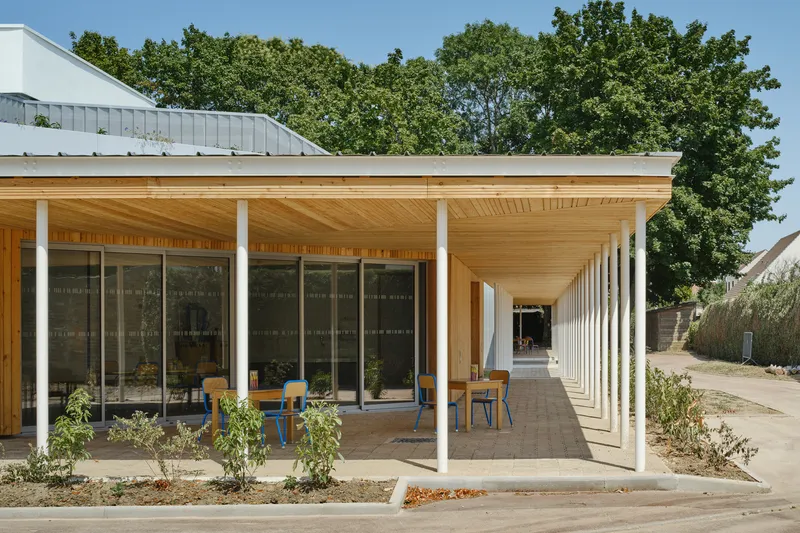 Covered walkway with timber soffit and white columns extending along a glazed facade under mature trees