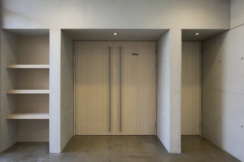 Entrance alcove with vertical wood door flanked by concrete walls and recessed shelving niche