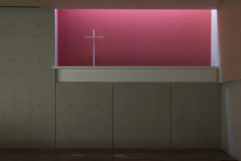 Interior chapel wall with high clerestory window revealing a pink-lit cross against the sky
