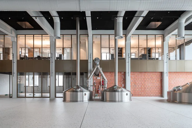 Double-height interior space with copper brewing kettles beneath glazed mezzanine offices and structural steel columns