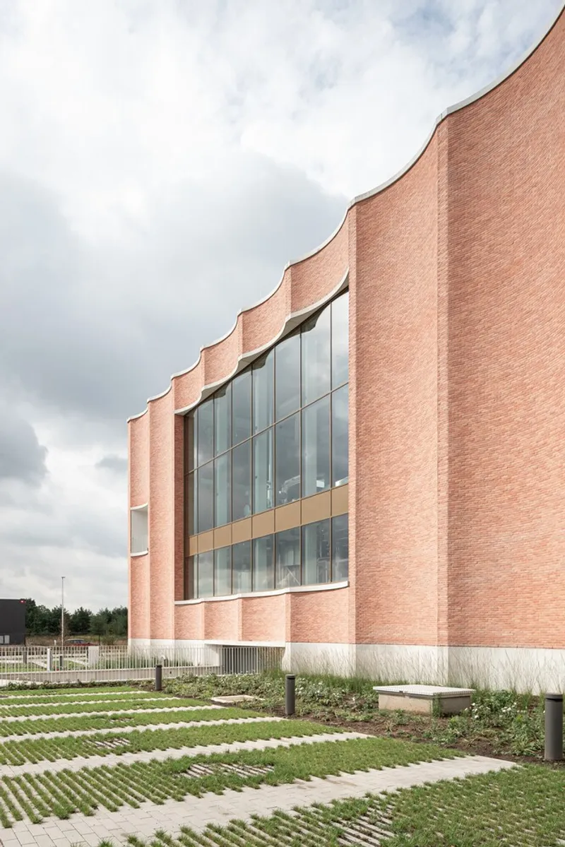 Pink brick facade with scalloped parapet and central glazed window above patterned paving under cloudy skies