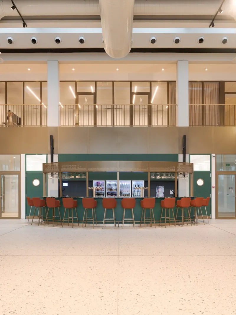 Green tile bar with orange stools beneath a glazed mezzanine level in the main atrium