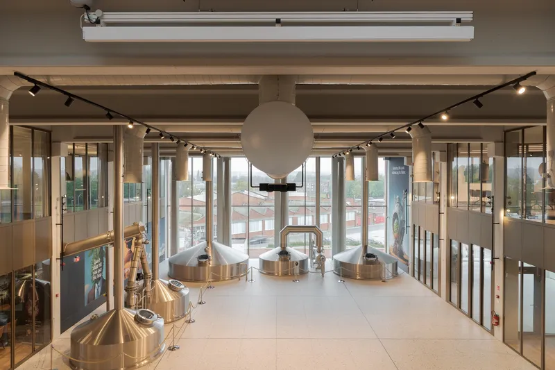 Copper brewing vessels behind glass partitions in double-height visitor gallery with track lighting and terrazzo flooring