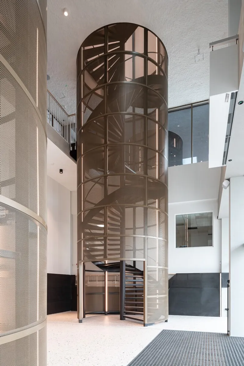 Bronze metal spiral staircase enclosed in cylindrical glass shaft within white painted multi-level interior