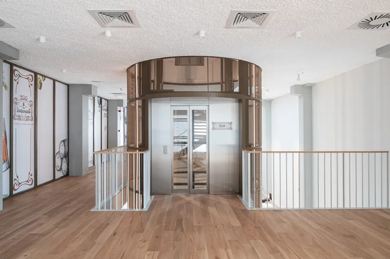 Bronze-clad cylindrical elevator core flanked by vertical metal railings on a timber floor under recessed ceiling lighting