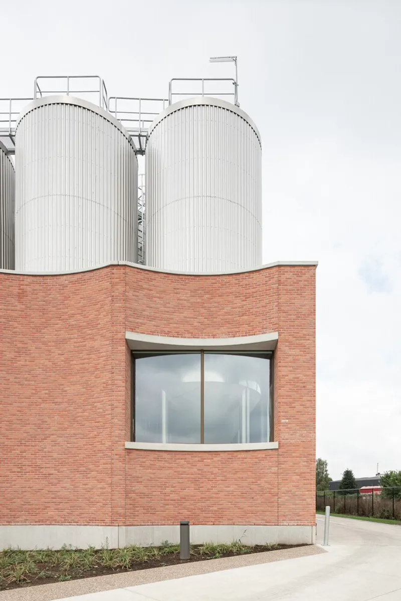 Curved glazed bay window set in a red brick facade beneath cylindrical metal silos and cloudy sky
