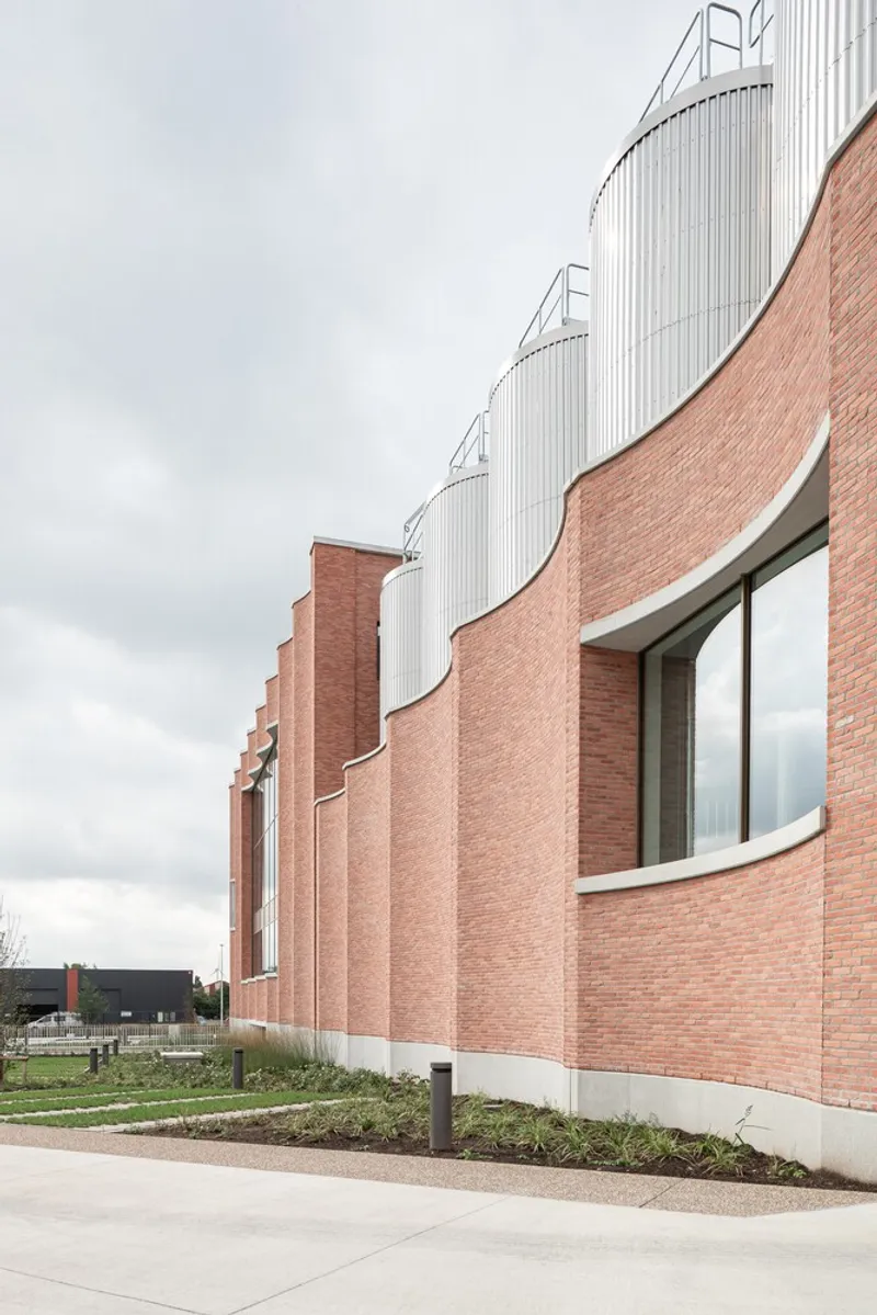 Curving red brick facade with tall windows and cylindrical white metal silos rising above under overcast skies