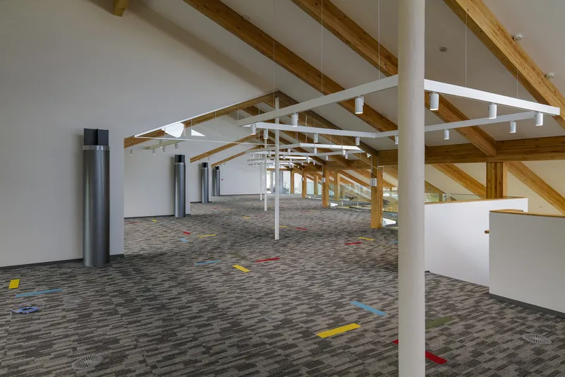 Interior hall with white steel column, suspended ring lights and timber post-and-beam structure under a vaulted ceiling