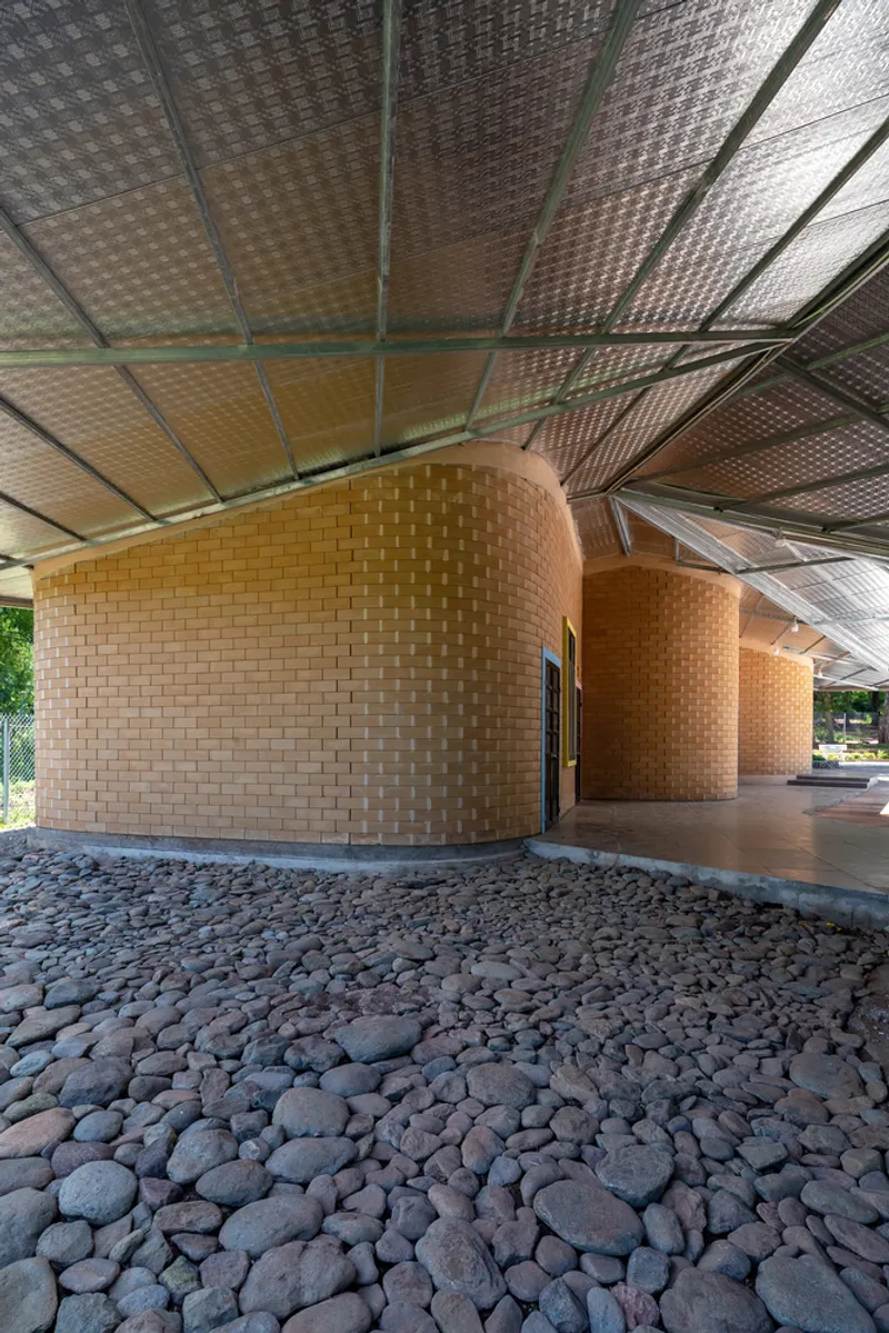 Three cylindrical woven brick volumes under a translucent paneled roof with cobblestone ground cover