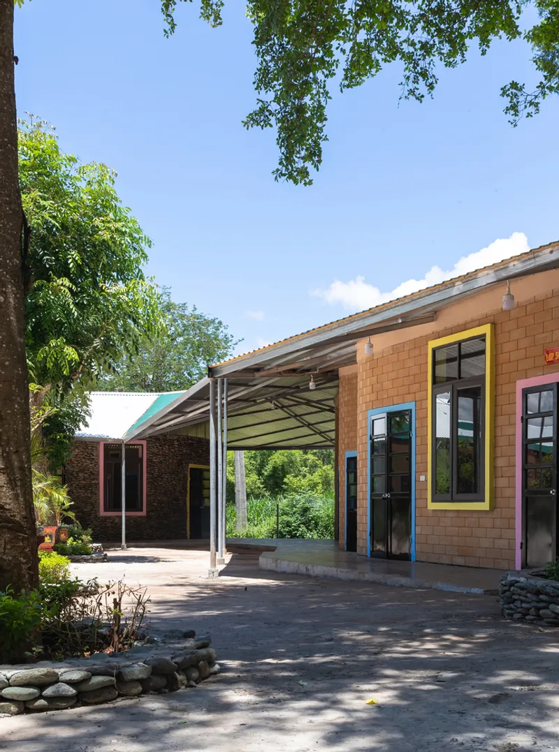 Covered walkway connecting brick and tile-clad volumes beneath overhanging corrugated roofs framed by trees
