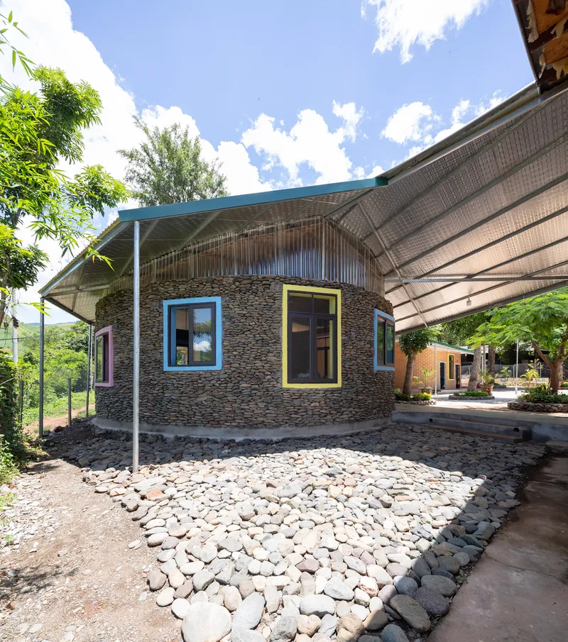 Curved stone wall with blue and yellow framed windows beneath a woven bamboo canopy on a sunny day