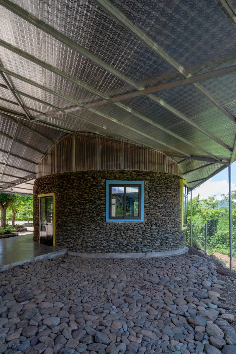 Curved stone wall with blue-framed window beneath a translucent corrugated roof over a cobble courtyard