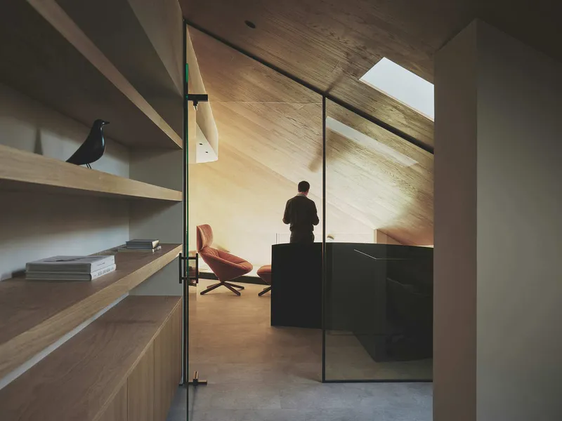 View through glass partition to study with figure standing at desk beneath angled timber ceiling and skylight