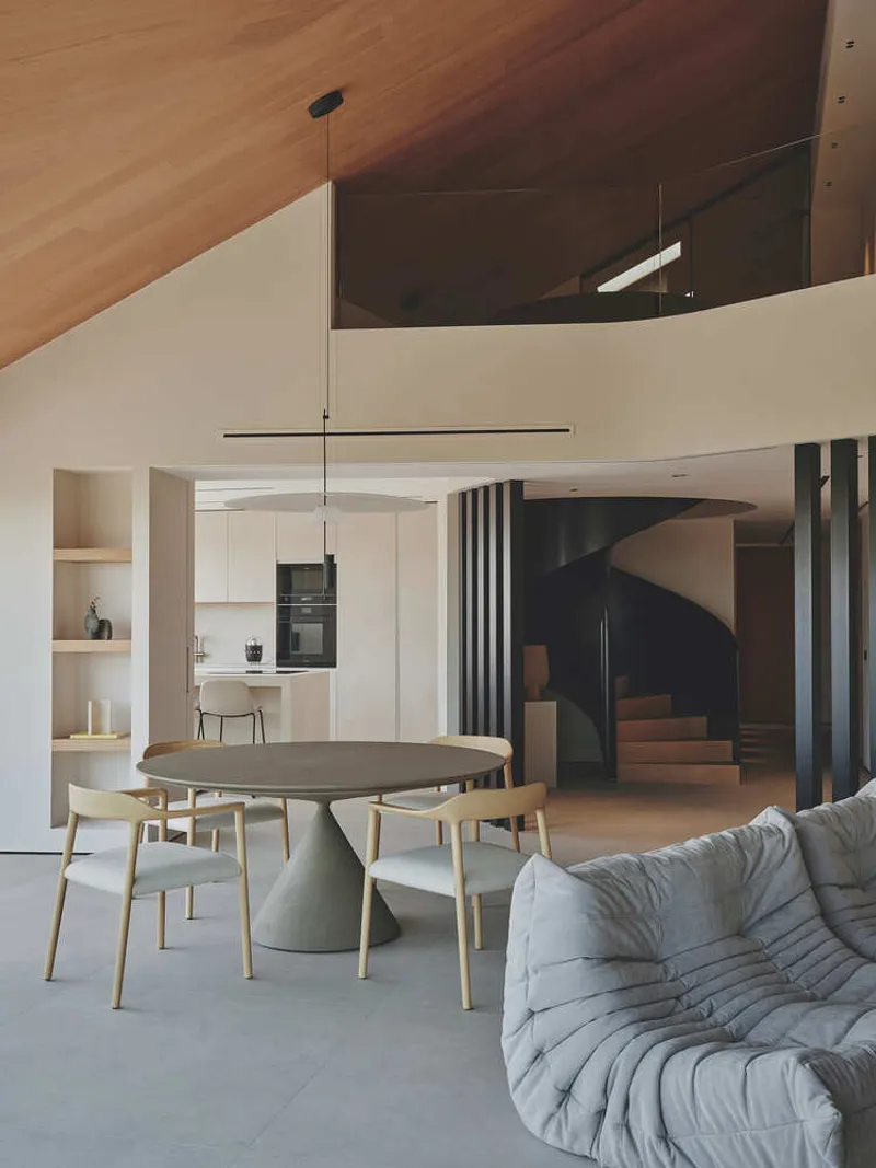 Dining area showing curved staircase enclosed by dark vertical slats beneath double-height ceiling volume