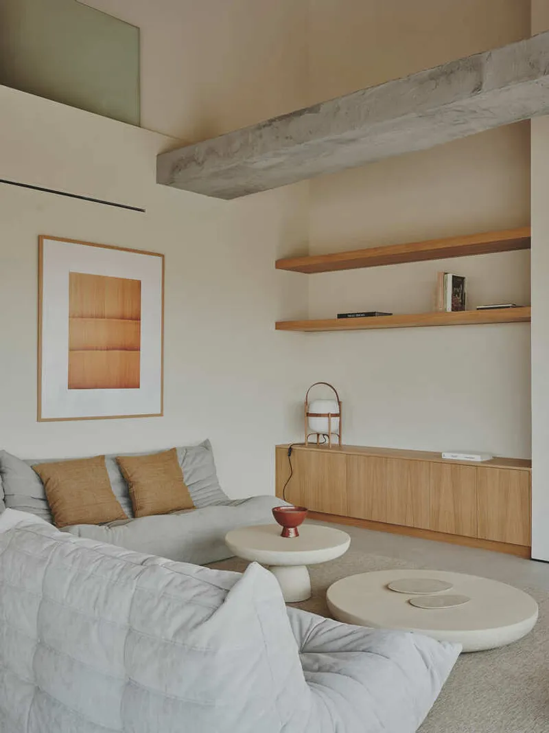 Living room with exposed concrete beam, timber shelving, and low circular coffee tables in soft daylight