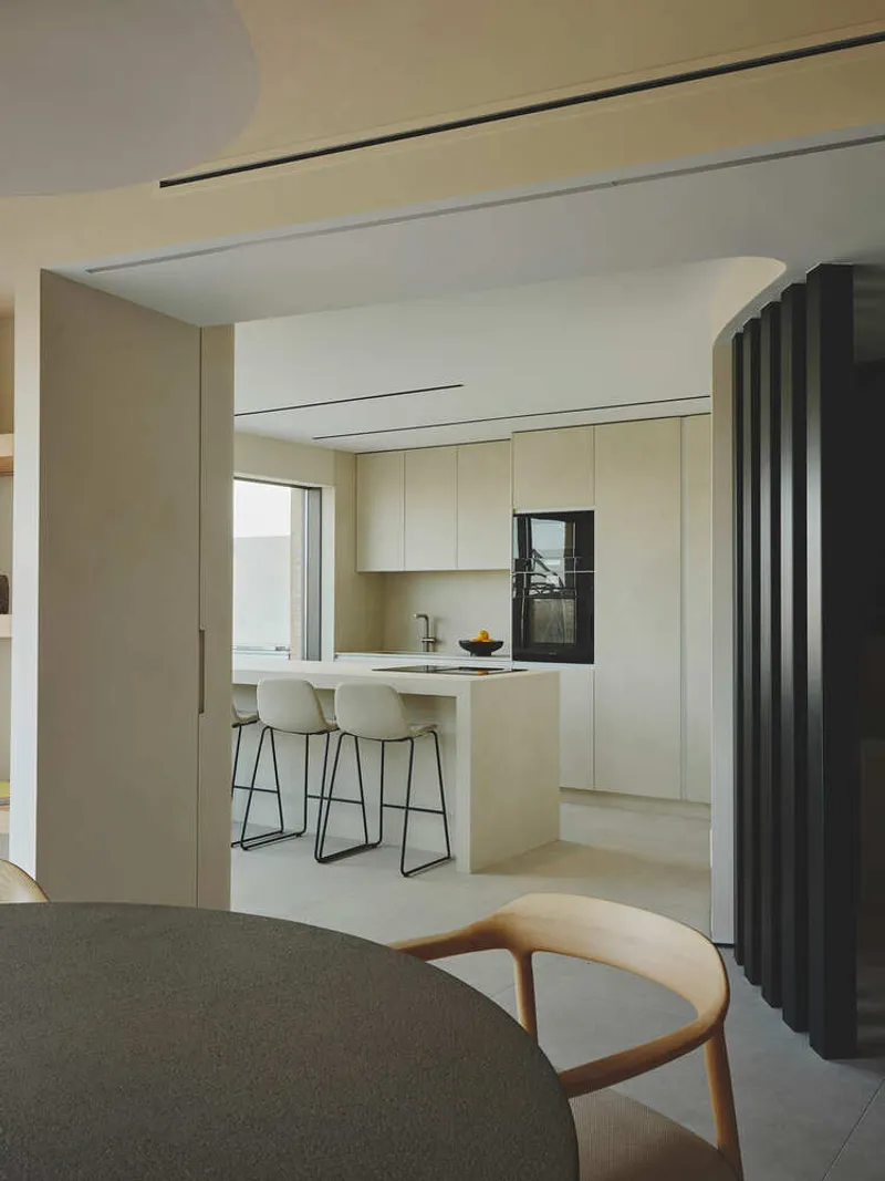 View through wide opening to kitchen island with pale wood cabinetry and white bar stools