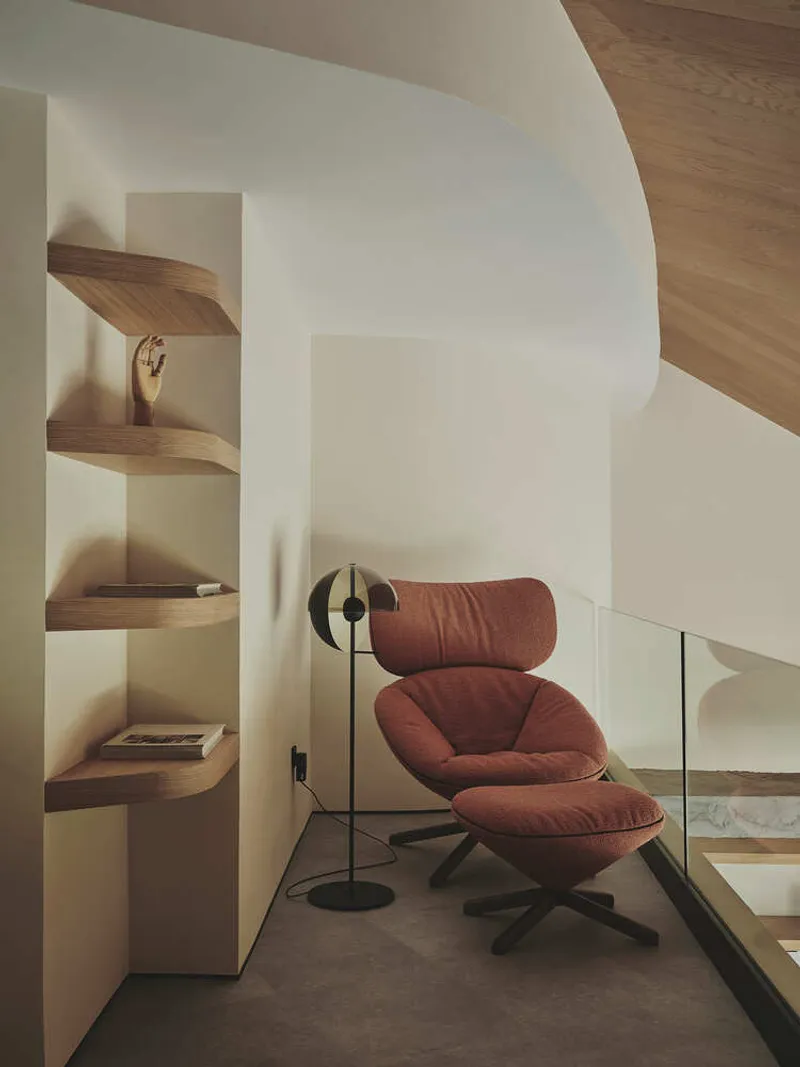 Reading nook with curved white wall, timber corner shelves, and rust-colored lounge chair by black floor lamp