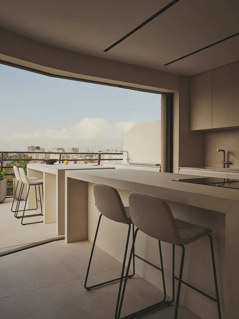 Kitchen counter with bar stools opening onto a terrace overlooking a low-rise urban skyline at dusk