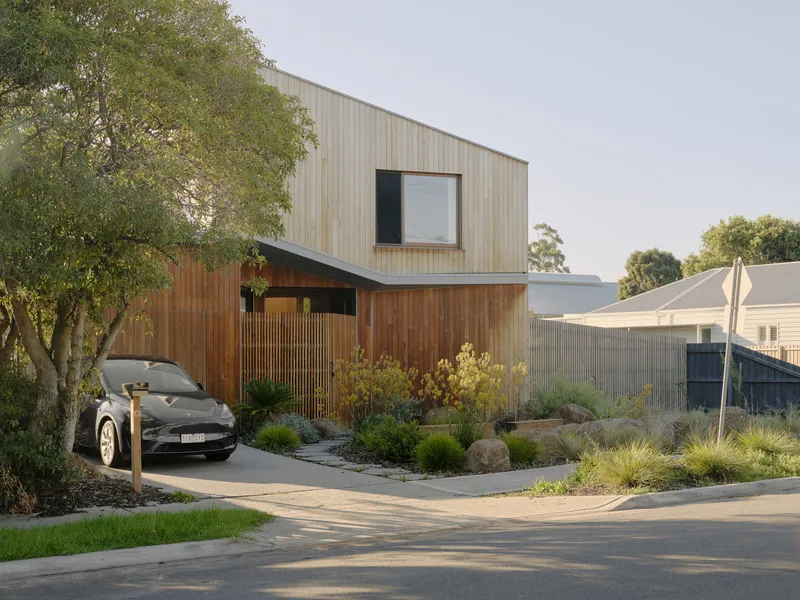 Street view of the two-storey timber-clad facade with native grasses and carport in afternoon light