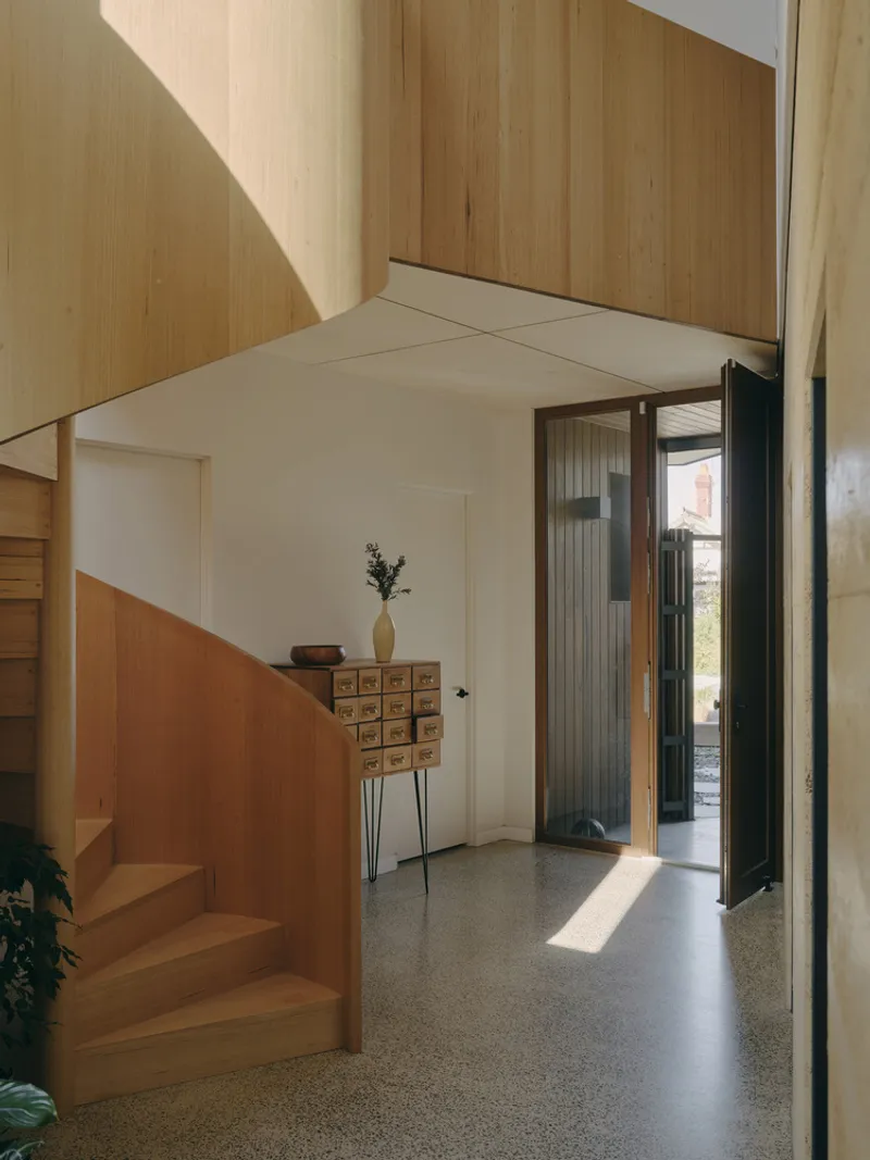 Entry hall with timber staircase and curved plywood ceiling catching sunlight from the open door