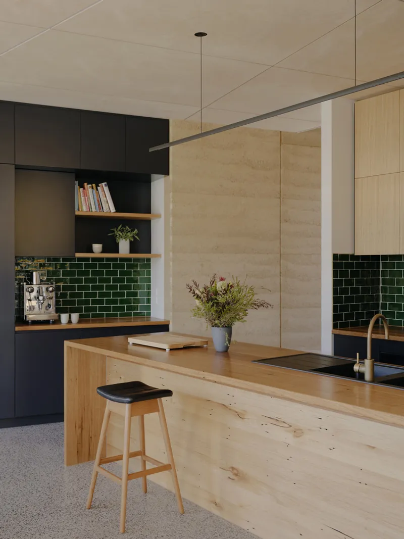 Kitchen island bench in raw timber with rammed earth wall and dark cabinetry beyond