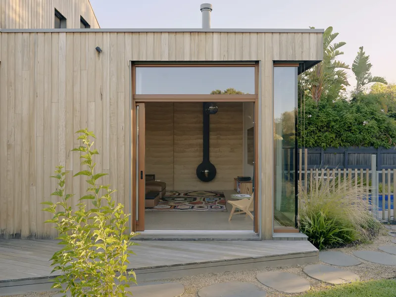 Vertical timber cladding with full-height glazed opening looking into rammed earth reading nook at dusk