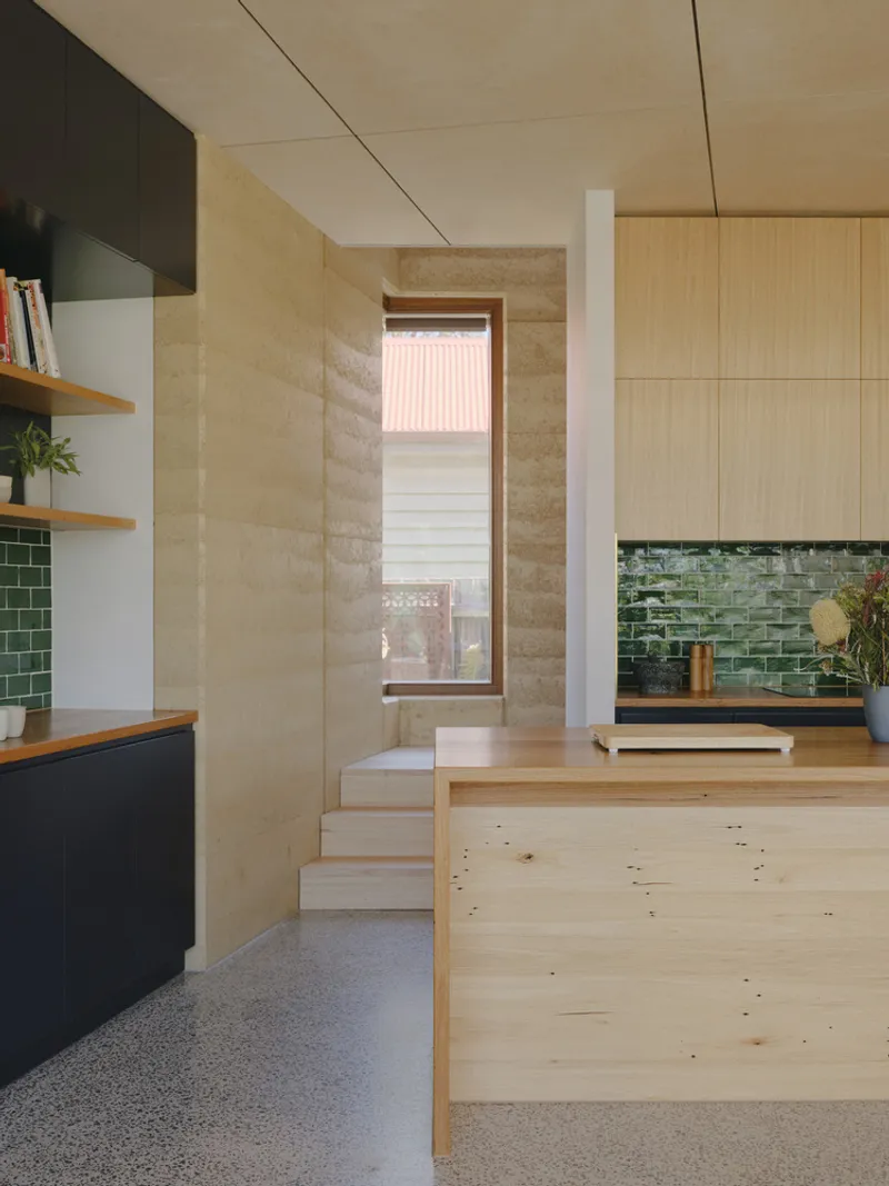 View through split-level kitchen to tall window framed by layered rammed earth walls