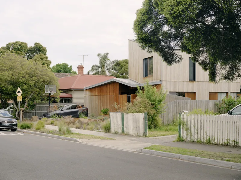 Street view of vertical timber clad volumes behind a picket fence and native planting