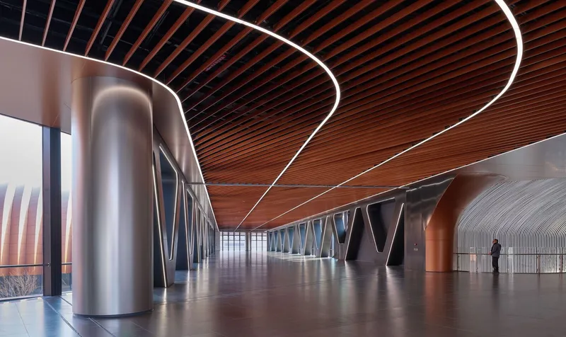 Interior corridor with curving timber slat ceiling, sculptural steel columns and a lone figure at the far end