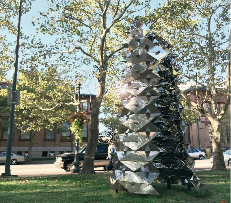 Mirrored sculptural tower standing in a tree-lined park on an autumn afternoon