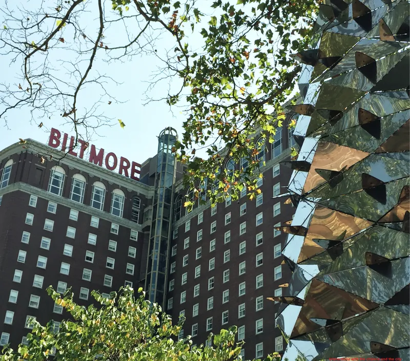 Reflective metal panel tower with faceted surfaces beneath overhanging tree branches in daylight