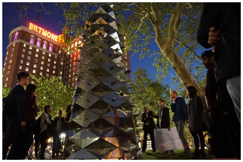 Twisting mirrored metal tower installation in a public plaza surrounded by visitors at twilight