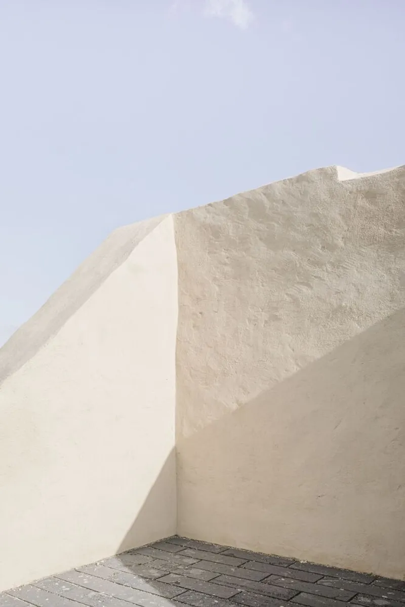 Corner detail of plastered walls meeting at sharp angles above a dark paved floor under pale sky