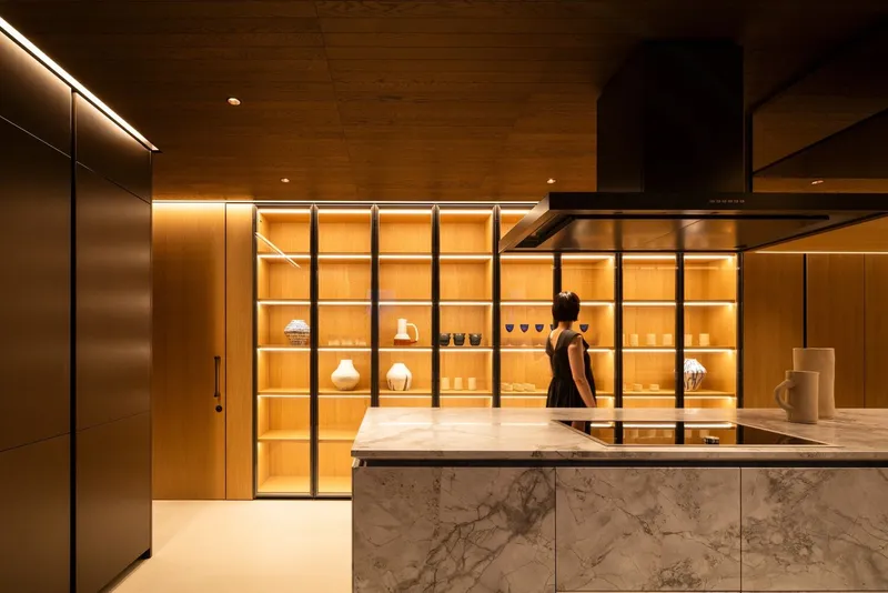 Kitchen island clad in veined marble with backlit glazed display shelving and person standing beyond