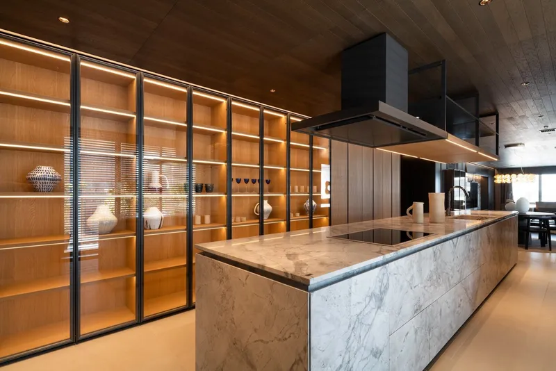 Marble kitchen island with black range hood and illuminated glass-fronted oak cabinetry along the wall